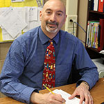 Joe Prestiani wearing a blue shirt and red tie sitting at his desk writing on a piece of paper and smiling.