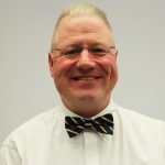 A man wearing a white shirt, striped bow tie, glasses, smiling broadly