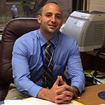 Assistant Principal Seth Siegel wearing a blue shirt and striped tie sitting at his desk