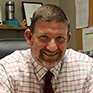 A man with short light brown hair, facial hair on his chin and mustache smiles. He is wearing a check collared shirt and a maroon tie.
