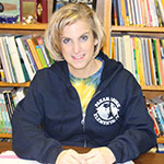 Colleen Delles smiling, sitting at her desk with book cases behind her; chin-length blonde hair, wearing a Pine Bush zippered sweatshirt