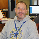 Brian Breheny smiling, sitting at his desk and wearing a Pine Bush Spirit hooded sweatshirt