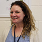 A woman with curly hair smiles. She is wearing a beige sweater and blue print dress and a blue lanyard around her neck.