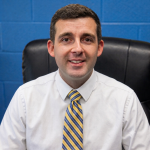A man wearing a white shirt and blue and gold tie sits in a chair and smiles. He has short dark hair.