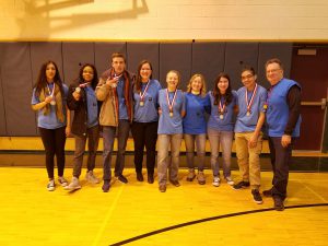Seven members of the Pine Bush High School Odyssey of the Mind team smile, link arms with their teammates and coaches, while wearing their medals for winning the regional competition.