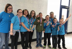 Seven members of the Pine Bush Elementary Odyssey team stand holding their trophy with their medals around their necks on red, white and blue ribbons. Their coaches are with them. The boy on the right has his arm extended high/