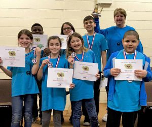 Seven members of the Pakanasink Odyssey of the Mind team, dressed in their royal blue shirts, hold their certificates and medals after winning the regional competition. Their coach holds the trophy up.