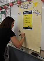 Teacher is the first to sign the No Place For Hate pledge. She is using a large black marker on yellow paper hung on the wall.