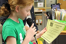 A young student in a bright green shirt holds the microphone in one hand and the No Place For Hate pledge in the other and recites it over the loud speaker for her school.