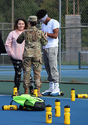 On a bright blue tennis court, one student is blindfolded as the Army recruiter dressed in camouflage explains what will happen to another student who will driect the blindfolded one.