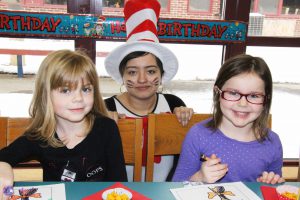 Two kindergarten girls sitting together smile for camera as they color a Dr. Seuss picture. Between them is a high school student dressed as the Cat in the Hat.