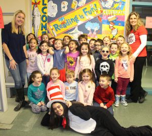 Twenty-two kindergarten kids are gathered with a high school student dressed in a black and white Cat in the Hat suit and red and white striped hat. Their two teachers flank them. All are laughing and smiling.