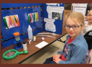 A young girl has her poster board with her science experiement with colorful strips and a magifying glass.