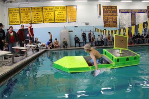 Young man in a tan camping hat paddles a neon green boat in the high school pool.