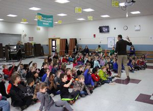 A large group of first grade students sit on the floor, many with their hands up.