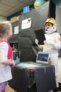 A fifth grade student is dressed as Neil Young in a space suite talling a girl his life story.
