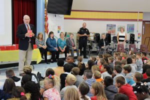 Third grade students sit on the cafeteria floor and listen to the Rotary respresentative who is holding a Webster's Dictionary in his hands.