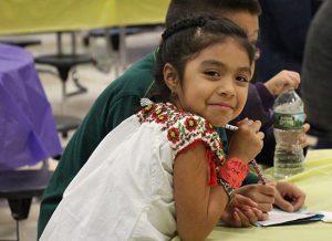 A little girl in a white shirt with red flowers smiling and coloring.