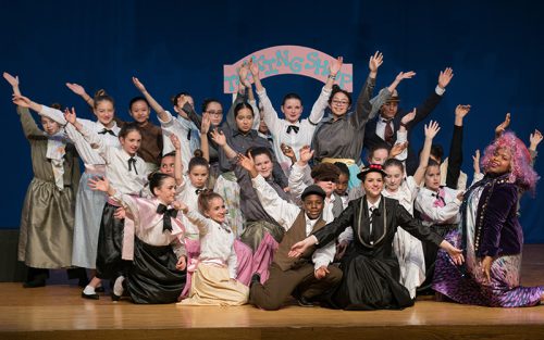 The cast of Mary Poppins in costumes with their arm reaching up