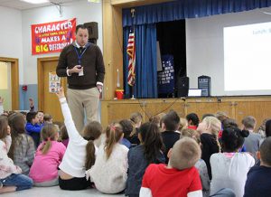 Assistant Principal talks to first graders who are sitting on the floor. One girl has her hand up.