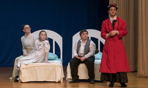 Mary Poppins dressed in a long red coat and hat, talks to the three children who are sitting on their beds.