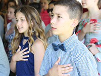 A fifth grader wearing a blue shirt and bow tie and a young girl with long blonde hair have their hands over their hearts as they recite the pledge