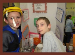 Two young students, one with a  clear shield over his face, in front of  their poster board.