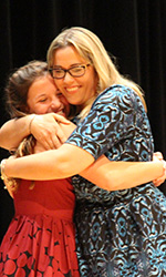 Fifth grade teacher with long blonde hair is smiling broadly wearing a blue dress hugs one of her students, wearing a red dress. 