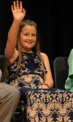 Fifth grade girl smiles and waves from the stage to her family 