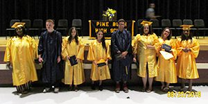 Six girls in gold graduation caps and gowns and two boys in black grduation caps and gowns stand in front of the Pine Bush podium .