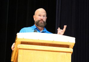 man with beard and blue shirt stands at the podium and speaks to the crowd