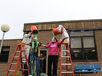 Standing on ladders, two jumpsuit-clad volunteers pour buckets of slime on two administrators