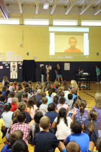 Students sit on the gym floor and watch a video of the meaning of No Place For Hate