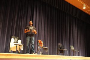 Man in jeans and black t shirt on stage acting alongside four empty chairs, which have different clothing on them.