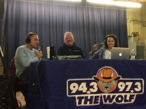 Radio hosts sit on either side of man being interviewed. Table has the Wolf radio logo