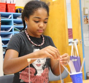 Student in black shirt and pink and black necklace builds a structure out of plastic straws