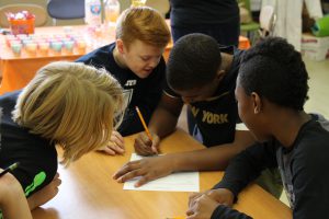 A group of four fifth-grade boys huddled together to work on their experiment. One is writing on a piece of paper.