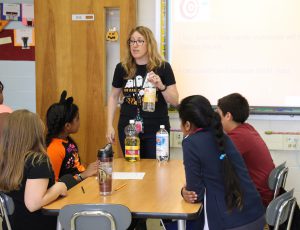 A teacher with blond hair holding a bottle of seltzer while talking to four fifth-grade students