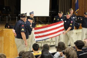 Men in their legion caps fold a flag in front of an auditorium filled with middle school students