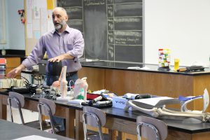Man with beard wearing a light colored button down shirt talks to students and has many medical props on a table in front of him.