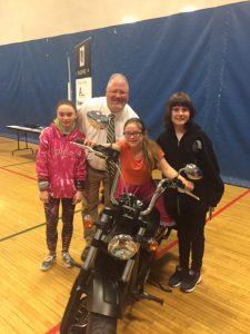 A man and woman pose with two middle school girls, one of which is sitting on a motorcycle in the gym.