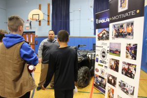 .A man wearing a quarter zip gray pullover jacket stands in front of a four-wheeler and talks to several students.