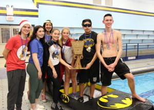 Seven high school students, two in Batman shirts, one dressed as Wonder Woman, stand near their boat and hold the Pine Bush boat race trophy, which is made of cardboard. The boat is black with a yellow and black batman symbol on it.