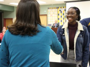 Woman with dark hair and a blue sweater shakes hands with a smiling young woman wearing a denim jacket.