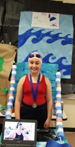 Girl in red bathing suit, swim cap, goggles and a gold medal around her neck smiles. Her desk has lane markers and looks like a swim lane.