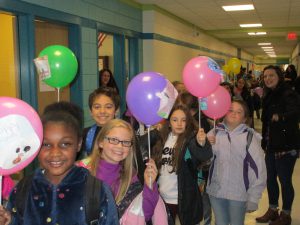 Many fourth-grade students in a line walking through a hallway holding up green, purple and pink balloons, all smiling.