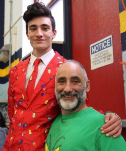 A high school student in a red suit with Christmas lights on it stands with a teacher in a green shirt. Both are smiling.