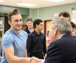 Young man with blonde hair smiling in blue t shirt while shaking hands with man in a blue suit jacket.