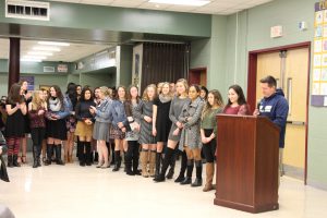 Approximately 20 middle and high school girls stand in a line next to a podium where their coach, a man with a blue hoodie talks into a microphone