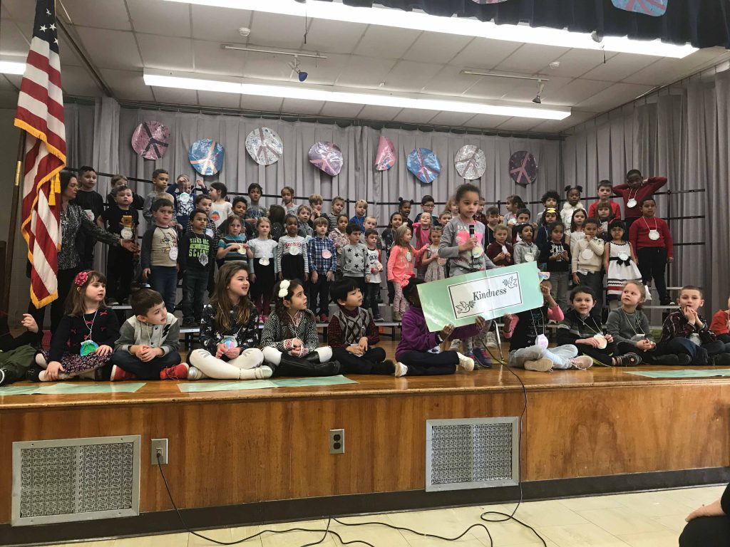 large group of kindergarten children on stage with peace signs above them and a girl holding a microphone and others holding a sign with the word "Kindness" on it.